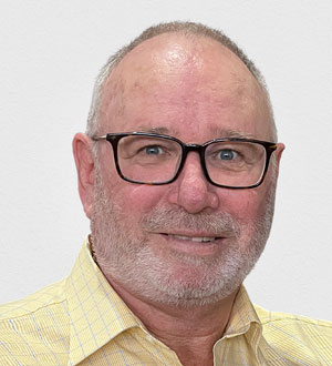 Alan Nicholls headshot wearing black frame reading glasses and yellow checkered shirt